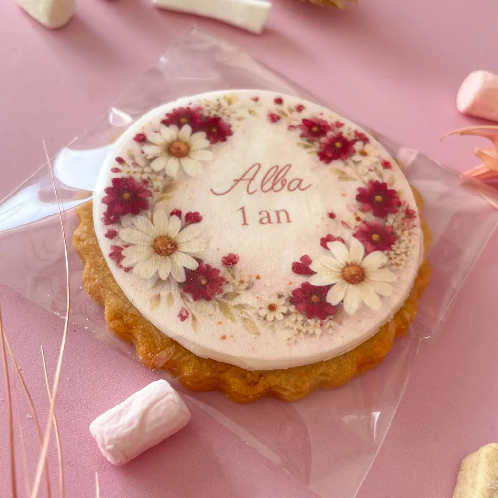 Un sablé rond « Alba 1 an » décoré d’une couronne de marguerites blanches et fuchsia, dans son sachet transparent sur un fond rose