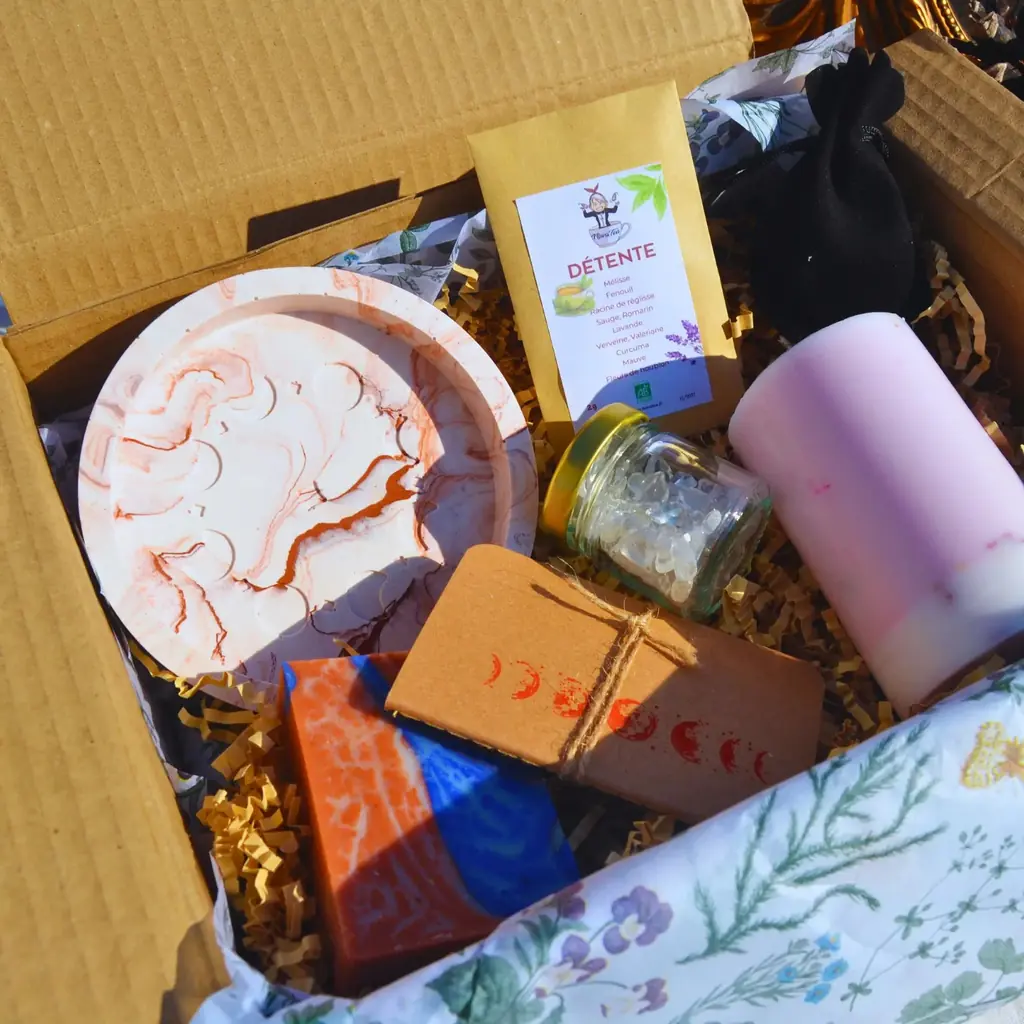 Un coffret cadeau ouvert dans sa boîte en carton : plateau en Jesmonite marbré, bougie pilier rose, savons, infusion et petit carnet en kraft