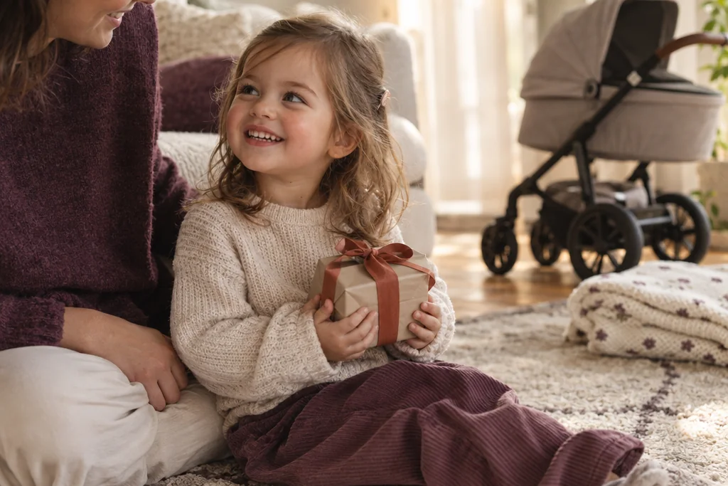 Une petite fille tient un cadeau emballé près d’un parent, devant une poussette, en attendant l’arrivée du bébé