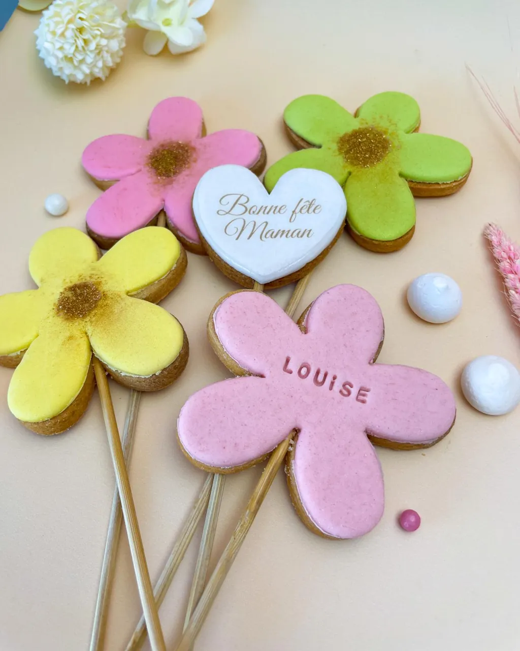 Des sablés en forme de fleurs roses, vertes et jaunes, dont un prénom gravé « Louise » et un cœur « Bonne fête Maman »