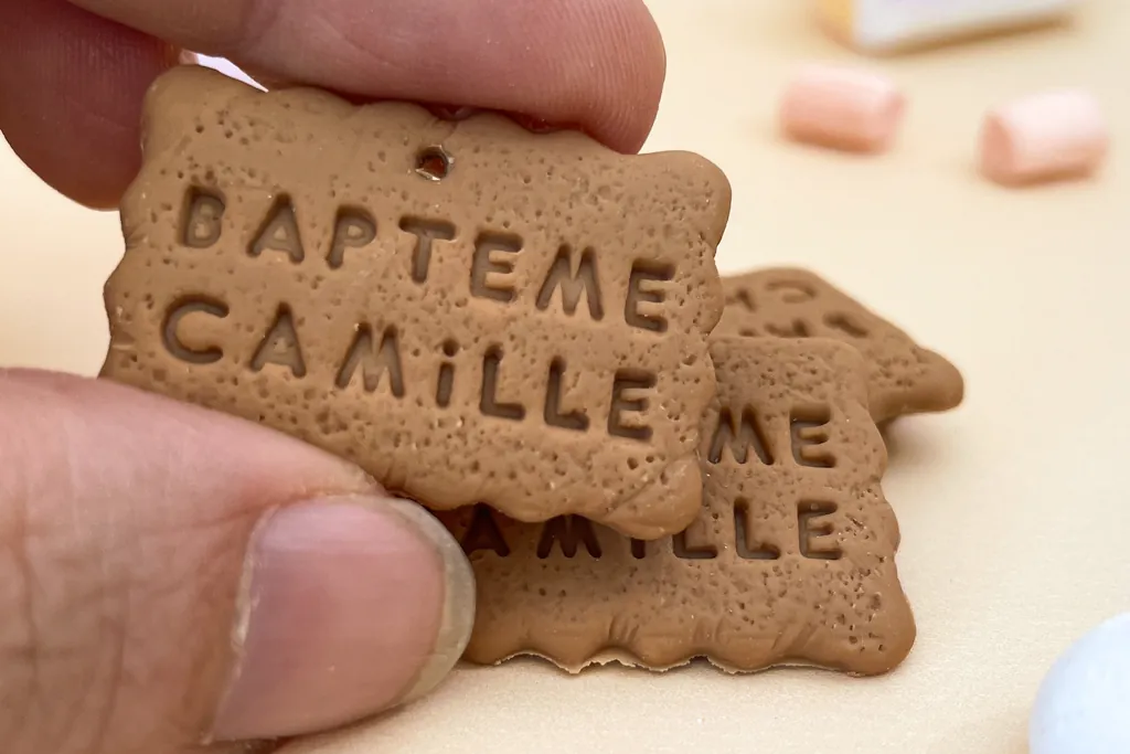 De petits biscuits en porcelaine froide en forme de petit-beurre, gravés « Baptême Camille », tenus entre deux doigts