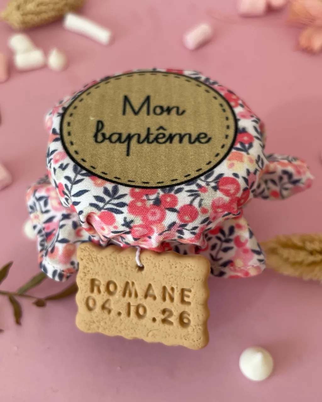 Un petit pot à dragées coiffé d’un tissu liberty à fleurs roses marqué « Mon baptême », avec une étiquette en forme de biscuit gravée « Romane 04.10.26 »