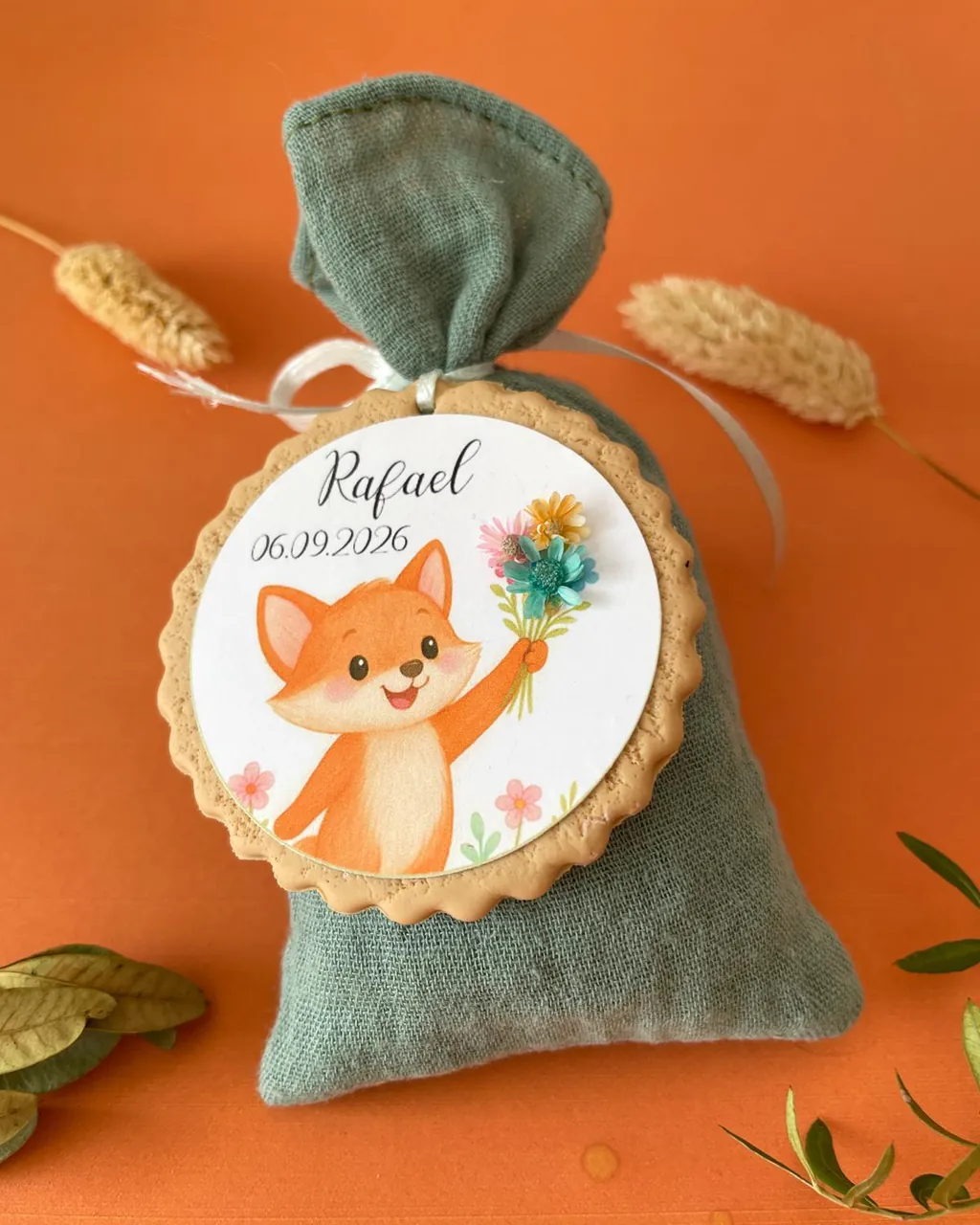 Un sachet à dragées en tissu vert sauge noué d’un ruban, orné d’un médaillon « Rafael 06.09.2026 » où un renardeau tend un bouquet de fleurs