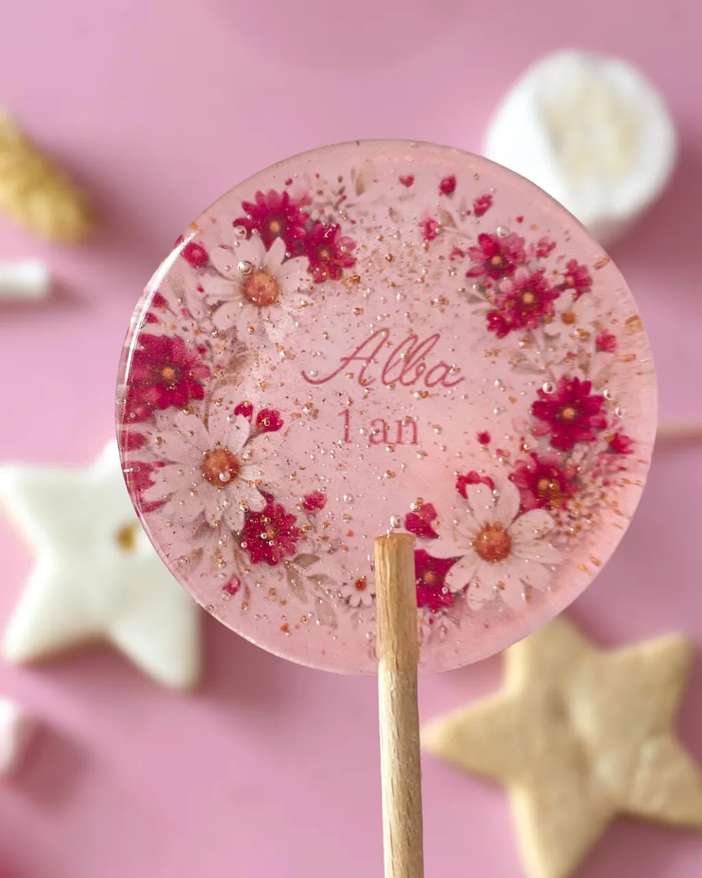 Une sucette ronde et transparente « Alba 1 an » décorée d’une couronne de marguerites blanches et fuchsia, devant des sablés en forme d’étoile