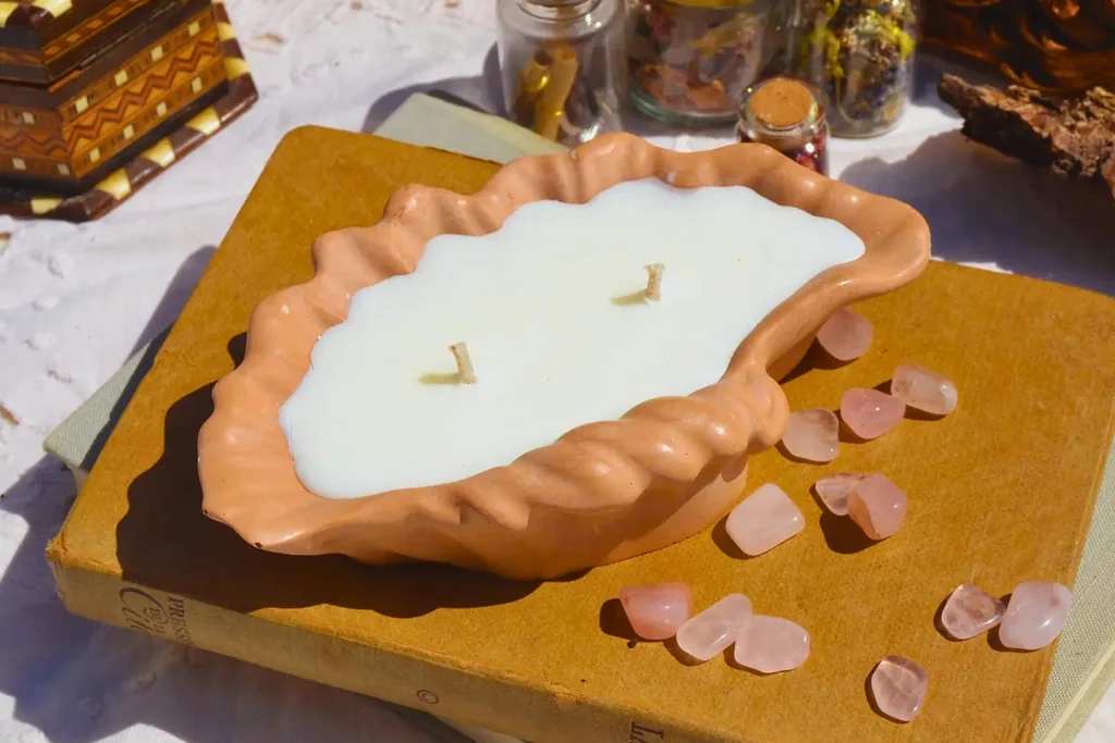 Une bougie végétale coulée dans un coquillage en Jesmonite terracotta, posée sur un livre ancien parmi des pierres roses