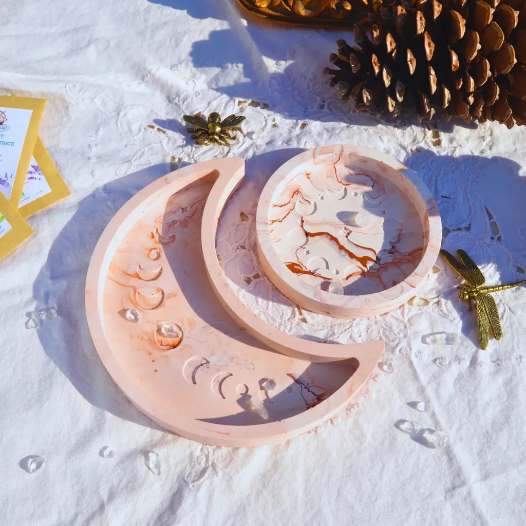 Deux plateaux en Jesmonite rose poudré, l’un en croissant de lune et l’autre rond, posés sur une nappe blanche près d’une pomme de pin