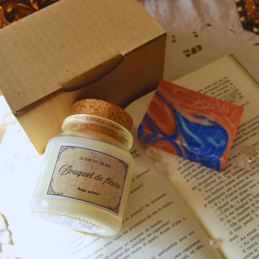 Une bougie végétale « Bouquet de fleurs » dans son pot en verre à bouchon de liège, à côté d’un savon marbré bleu et orange et de sa boîte en carton